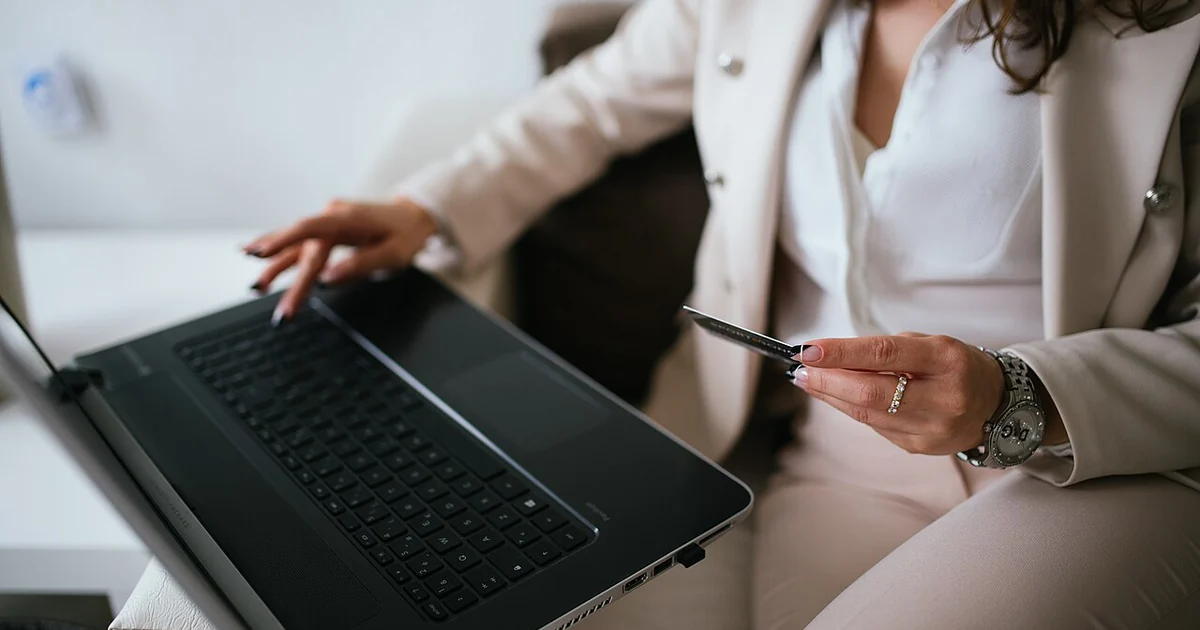 A business professional with curly hair dressed in a suit works on a laptop at home, holding a credit card in one hand and typing with the other, indicating an online shopping activity.